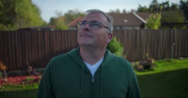 Arka bahçeye bakan bir adam ahşap çitlerin ve konut evlerinin yakınına günlük ve düşünceli bir an, banliyö mahallesi, yeşillik ve sakin bir çevre.