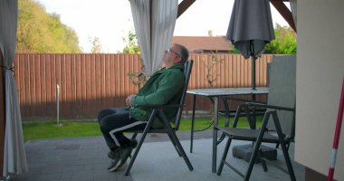 Bahçedeki örtülü pergola altında teras sandalyesine yaslanan adam, doğa ve huzurlu bahçe atmosferiyle çevrili huzurlu anın tadını çıkarıyor.