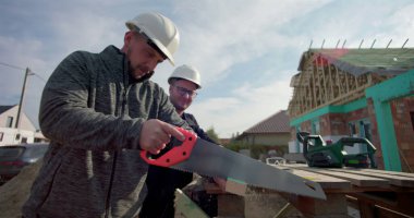 İnşaat işçisi ahşap tahtayı el testeresiyle kesiyor, iş arkadaşı gözlemliyor, inşaat sahasında takım çalışması, ev iskeleti görünür, hassasiyet ve işçiliğe odaklanmış.
