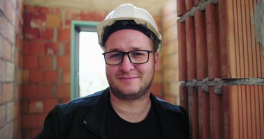 Tuğla duvara dayanmış inşaat işçisi, gülümsüyor ve güvenlik kaskı ve gözlüğü takıyor, tamamlanmamış bir binanın içinde, profesyonel varlığını ve güvenini gösteriyor.