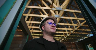 İnşaat işçisi açık çatı altında duruyor, başlık ve gözlük takıyor, inşaatın ilk aşamasında inşaat projesine odaklanıyor ve kararlılıkla bakıyor.