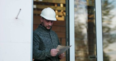 İnşaat işçisi kapıdaki planlar üzerinde çalışıyor, başlık takıyor, tamamlanmamış inşaat projesinin detaylarına odaklanıyor, inşaattaki hassasiyet ve kararlılığı aktarıyor.