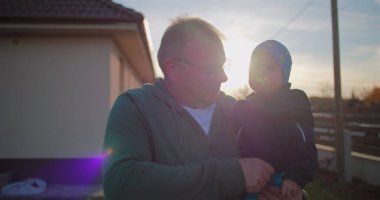 Baba, bebeğini emzik ile arka bahçede tutuyor. Çocuk kıyafetlerini nazikçe ayarlıyor. Huzurlu bir aile ortamında sevgi ve sevgi gösterisi yapıyor..