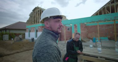 Ön planda güvenlik kaskı olan inşaat işçisi, gözlem alanı gelişimi, arka planda çalışan meslektaşı, kısmen inşa edilmiş ev yapısı görünür.