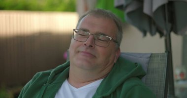 Avlu sandalyesinde uzanan, gözlüklü ve kapüşonlu bir adam. Nazikçe gülümseyen ve bahçede huzurlu bir anın tadını çıkaran bir adam.