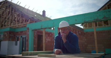 İnşaat işçisi inşaat alanında proje düşünüyor, eli çenesinde, ev yapısı ve arka planda çerçevesi olan düşünceli bir erkek inşaat işçisi.