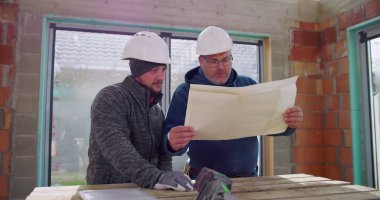 Bitmemiş binadaki ahşap masanın detaylı planlarını inceleyen inşaat işçileri, projeyi tartışıyorlar, güvenlik kaskı takıyorlar, takım çalışması yapıyorlar, işbirliği yapıyorlar.