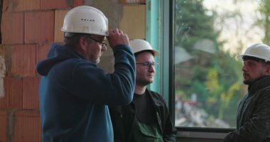 Cam kenarında sohbet eden inşaat işçileri, rahat ve günlük etkileşimler, kapalı alanda yenileme alanı, dostluk ve takım çalışması, güvenlik teçhizatı ve sert şapkalar.