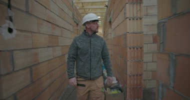 İnşaat işçisi miğfer takıyor, tamamlanmamış evin dar koridorlarında yürüyor, inşaat alanındaki tuğla duvarları ve tavan kirişlerini geçiyor.