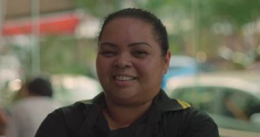 Kollarını kavuşturup güvenle gülümseyen Latin garsonun yakın plan portresi, arka planda yumuşak pencere ışığıyla restoranın içinde duruyor.