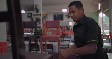 Genç Latin Amerikalı adam restoran vardiyasının başlangıcında sandalyeyi masadan kaldırıyor. Günlük yemek ortamında günlük kurulum rutinine başlıyor.