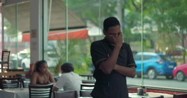 Restoran çalışanı, elleri gözünün üzerinde duruyor, vardiya sırasında yorgunluk ya da stres gösteriyor, arka planda görünen kapalı yemek alanı pencerenin yanında.