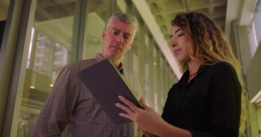 İş arkadaşları koridorda birlikte duruyor, tablet hakkındaki bilgileri inceliyor, profesyonel çevrede işbirliği yapıyor, dikkatle dinliyor.