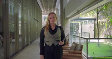 Modern koridorda elinde tabletle dikilen kadın, kameraya objektif bir ifadeyle bakıyor, profesyonel görev ya da buluşma için hazırlanıyor.