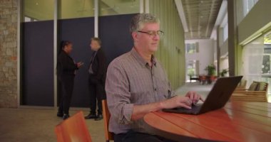 Modern çalışma ortamında dizüstü bilgisayarda yazan, arka planda konuşan, büyük pencerenin yanındaki ahşap masada oturan, profesyonel ortamda çalışmaya odaklanan bir adam.