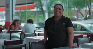 Latin garson sıcak bir gülümseme ile yemek alanında duruyor, rahat restoran ortamında koltukta dinleniyor ve yumuşak arka planda konuklar var.