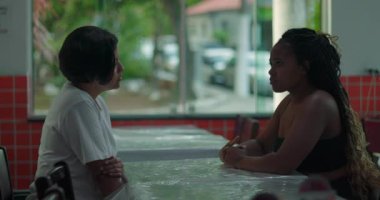 Lokanta masasında oturmuş iki kadın hararetli bir sohbete girişmiş, biri dikkatle dinlerken, diğeri arka planda yemek yerken.