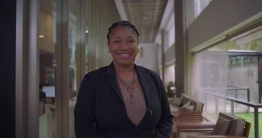 Afro-Amerikalı kadın şirket koridorunda güvenle gülümsüyor. İş ortamında dostane ve profesyonel bir ifade.