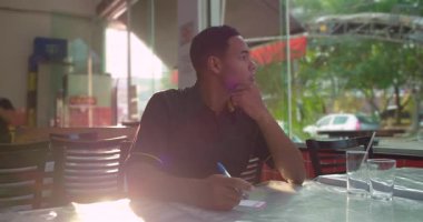 Afrika kökenli düşünceli Latin garson elinde kalem ve sipariş defteriyle duraklıyor, güneş ışığı restoranın içine doğru akarken pencereden dışarı bakıyor.