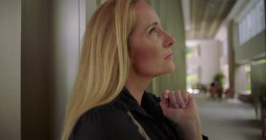 Yüksek tavan ve pencereli, parlak ışıklandırılmış kurumsal koridorda duvara yaslanırken düşünceli bir ifadeyle yukarıya bakan bir kadın.