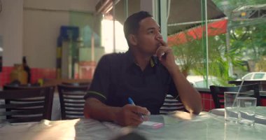 Afrika kökenli Latin garson, restoran masasında oturmuş elinde kalem ve sipariş defterini tutarak ayin başlamadan önce pencereden dışarı bakıyor.