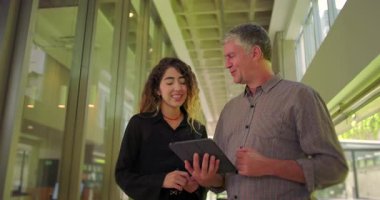 Kadın ve erkek tablet tutarken gülümsüyor, modern ofis koridorunda işbirliği yapan takım alışverişi, rahat ve verimli bir tartışmayla meşgul.