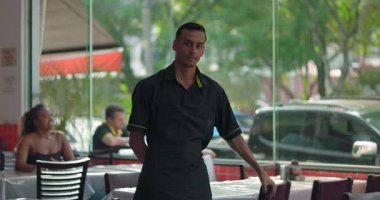 Genç restoran çalışanı masa ve cam pencerenin yanında poz veriyor, tarafsız bir ifadeyle sandalyeye yaslanıyor, arka planda oturan müşteri, görünür kentsel ortam