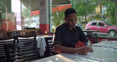 Odaklanmış Afrika kökenli Latin garson, restoran masasındaki sipariş defterine yazıyor. Konsantrasyon içinde baş aşağı, büyük pencerelerden gün ışığı süzüyor.