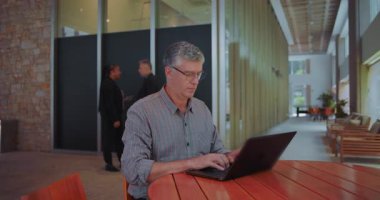 Orta yaşlı bir adam ortak ofis alanında dizüstü bilgisayarla çalışmaya devam ediyor, arka planda konuşan iş arkadaşlarıyla odaklanıyor, endişelerin üstesinden geliyor ve üretken kalıyor.