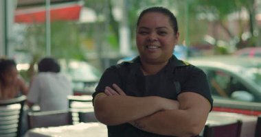 Önünde kolları bağlı bir şekilde gülümseyen Latin garson. Lokanta yemek odasında duruyor. Arka planda hafif gün ışığı ve masalar var.