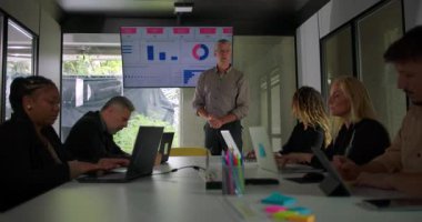 Yönetici olarak toplantıya katılan çeşitli profesyoneller ekibi, parlak ofis konferans odasında veri grafiklerini gösteren sunum ekranına işaret ediyor