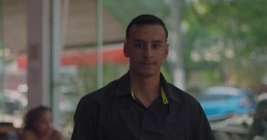 Restoranın içindeki genç erkek garsonun yakın çekimi, kameraya güpegündüz bakıyor ve pencereden dışarı park edilmiş arabaları görüyor.