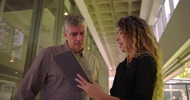 Ekip, koridordaki tablet içeriğini inceliyor, kadın fikirleri sunarken erkek dikkatle izliyor, ofis ortamında profesyonel işbirliği yapıyor.