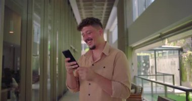 Genç adam telefonu tutuyor ve ekranda gülümsüyor. Profesyonel ofis koridorunda neşeli anlar yaşıyor. Sıradan ve dostane bir atmosfer.