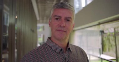 Orta yaşlı bir adam, yakından çekilmiş bir portrede, şirket koridorundaki kameraya bakıyor. Kendine güvenen ve odaklanmış bir ifade, olgun bir profesyonel görünüm.