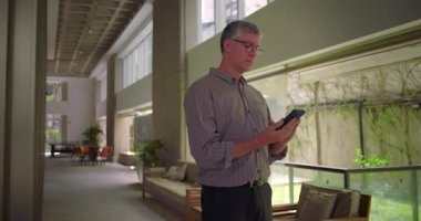 Orta yaşlı bir adam ofis koridorunda duruyor cep telefonu kullanıyor ve modern iş ortamında mobil cihazlarla etkileşime geçiyor.