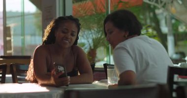 Gülümseyen kadınlar restoran masasında oturup akıllı telefonlara bakıyorlar. Parlak şehir yemekhanesinde gülüşlerini paylaşıyorlar.