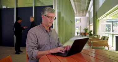 Modern açık uzay ofisindeki dizüstü bilgisayarda çalışan adam, büyük pencerelerin yanındaki ahşap yuvarlak masada oturuyor, odaklanmış ve daktilo ediyor, doğal elementlerle profesyonel ayarlar yapıyor.