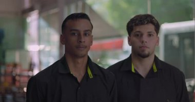 Yan yana duran iki erkek restoran çalışanı, ciddi ifadeler, büyük pencereli ve arka planda sokak manzaralı siyah üniformalar giyiyorlar.