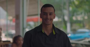 Pencerenin dışındaki bulanık şehir sokağında kameraya poz veren gülümseyen erkek restoran çalışanı, kendine güvenen ifade ve günlük ortam