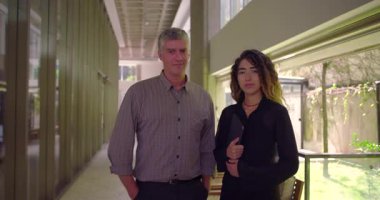 Ofis koridorunda yan yana duran iki profesyonel, tablet tutan ve kameraya bakan kadın, elleri cepte olan adam, her ikisi de kendine güvenen ifadeler sergiliyor.