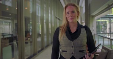Koridorda elinde tabletle duran bir kadın, kendinden emin ve dostane bir ifade takınıyor, modern çevrede liderlik, açıklık ve profesyonel bir varlık öneriyor.