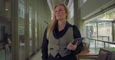 Modern koridorda elinde tabletle duran kadın, profesyonelliği ve kendine güveni yansıtırken hafif bir gülümseme ve ileriye doğru bakışla duruyor.