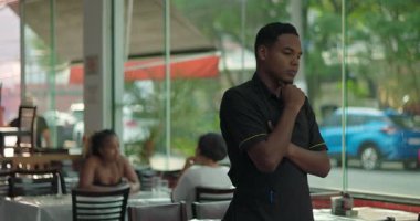 Düşünceli bir ifadeyle pencerenin yanında duran genç erkek restoran çalışanı, elleri çenesinde, yemek alanında sakin bir anda yansıtıcı gibi görünüyor.