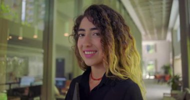 Özgüvenli bir ifadeyle gülümseyen genç bir kadın modern ofis koridorunda cam duvarın yanında duruyor tableti tutuyor ve kameraya bakıyor.