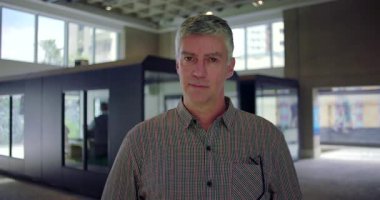 Orta yaşlı bir adam ofis koridorunda dikilip arka planda nötr bir ifadeyle modern profesyonel çalışma alanına bakıyor.