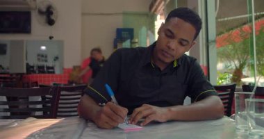 Latin Amerikalı genç bir adam restoran vardiyası sırasında not defterine yazıyor. Yemek masasında otururken ifadesine odaklanmış.