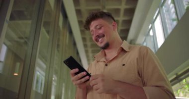 Ofis koridorunda akıllı telefona gülümseyen genç adam, mobil cihaz kullanırken neşeyle tepki veriyor, modern iş ortamında rahat ve neşeli bir an.