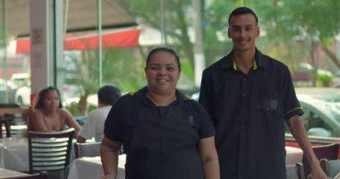 Latin garson ve garson yemek odasında yan yana dururken kameraya gülümsüyor. Arka planda açık renkli bir restoran var.