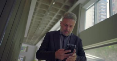 Orta yaşlı işadamı telefonu kontrol ederken hayal kırıklığı yaşıyor. Parlak modern ofis koridorunda haber veya mesajla vurgulanıyor.